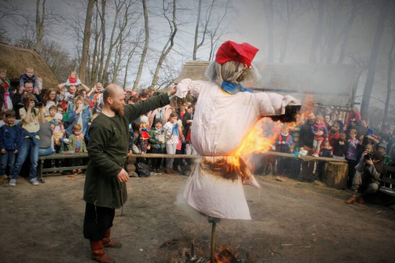 Mężczyzna w średniowiecznym stroju trzyma wysoką, płonącą kukłę marzanny. W tle tłum dzieci z dorosłymi. Parę osób robi zdjęcia.