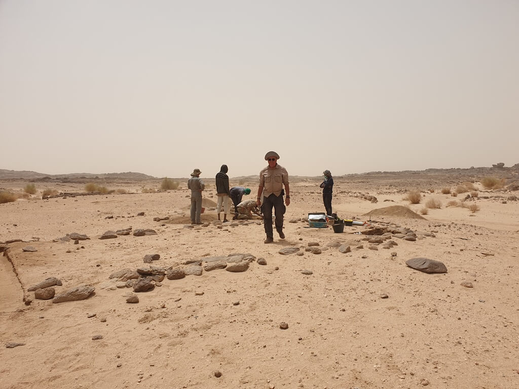 Fotografia: grupa pracowników podczas badań archeologicznych na pustyni. Na ziemi widać kamienne relikty (pozostałości) dawnej osady.