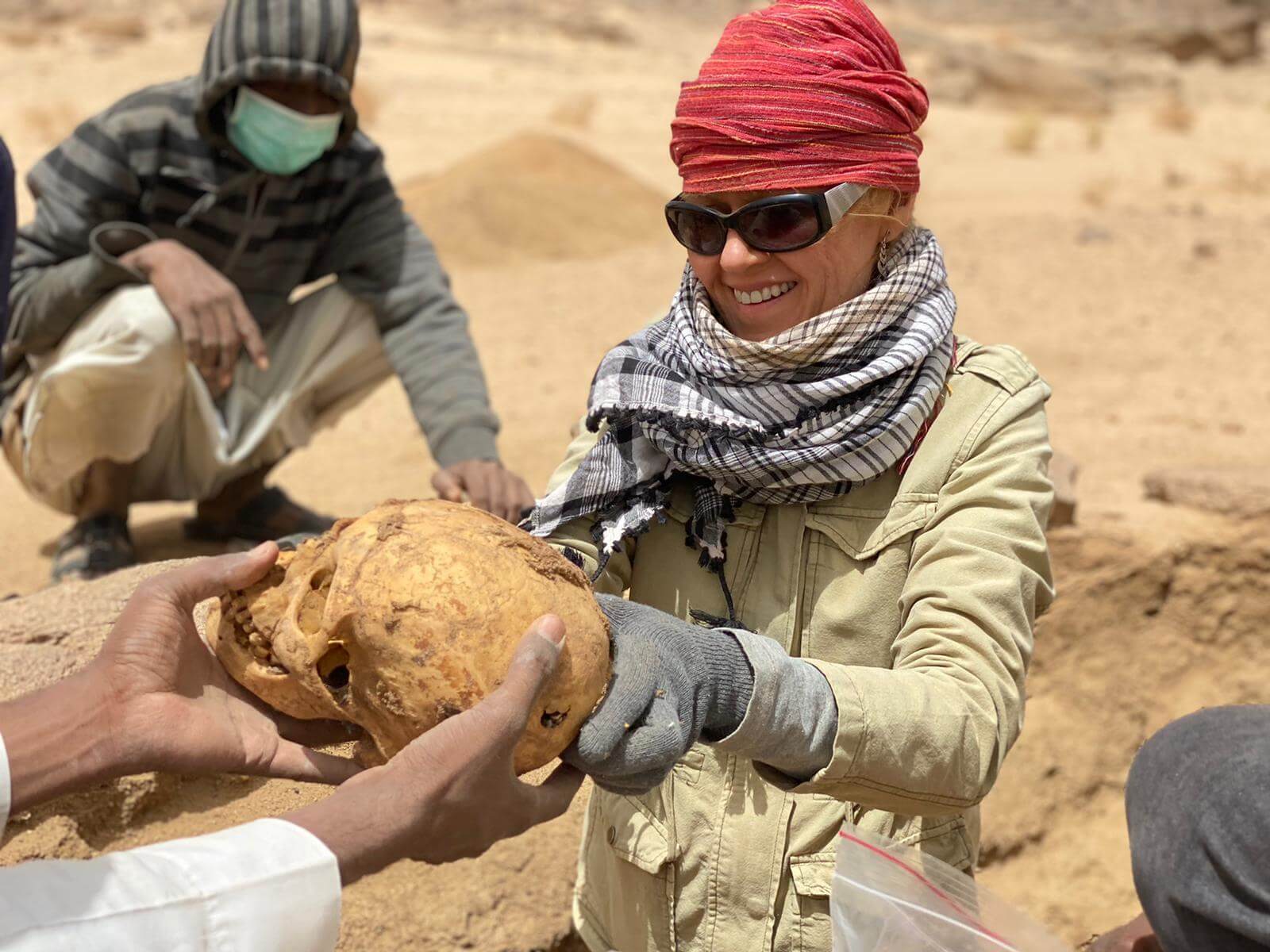 Fotografia: antropolożka w czerwonej chuście na głowie podaje komuś spoza kadru ludzką czaszkę odkrytą na pustyni.