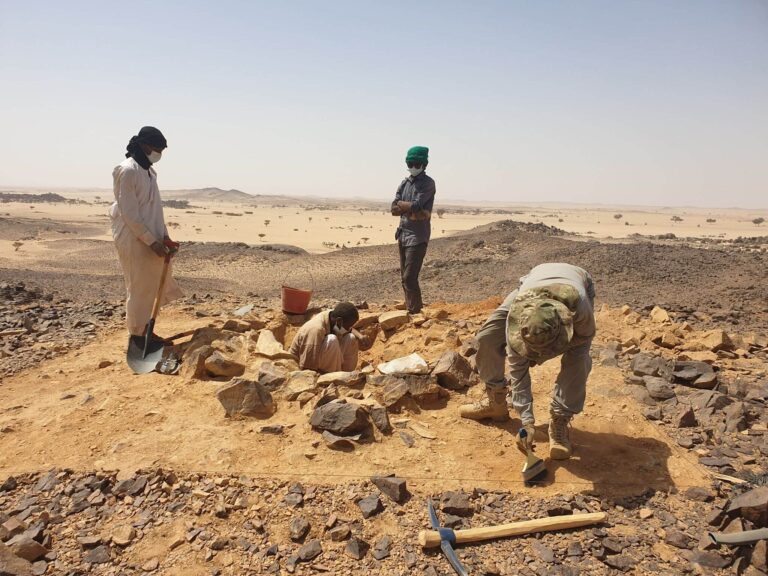 Fotografia: badania archeologiczne na pustyni. Archeolog odsłania obiekt (prostokątny zarys grobu) przy użyciu pędzla. Obok trzech tubylców z narzędziami.