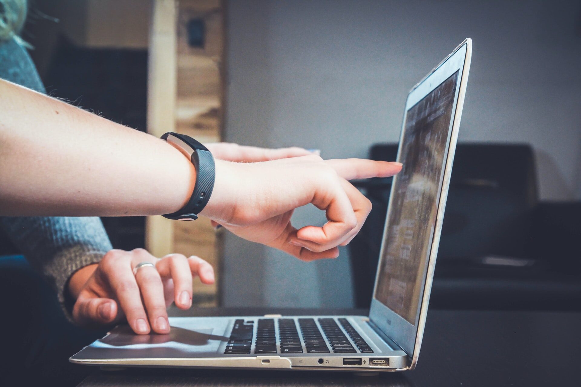 Zdjęcie przedstawia laptop oraz dłonie należące do dwóch różnych osób. Jedna dłoń przybliża się do ekranu komputera, wskazując coś palcem.