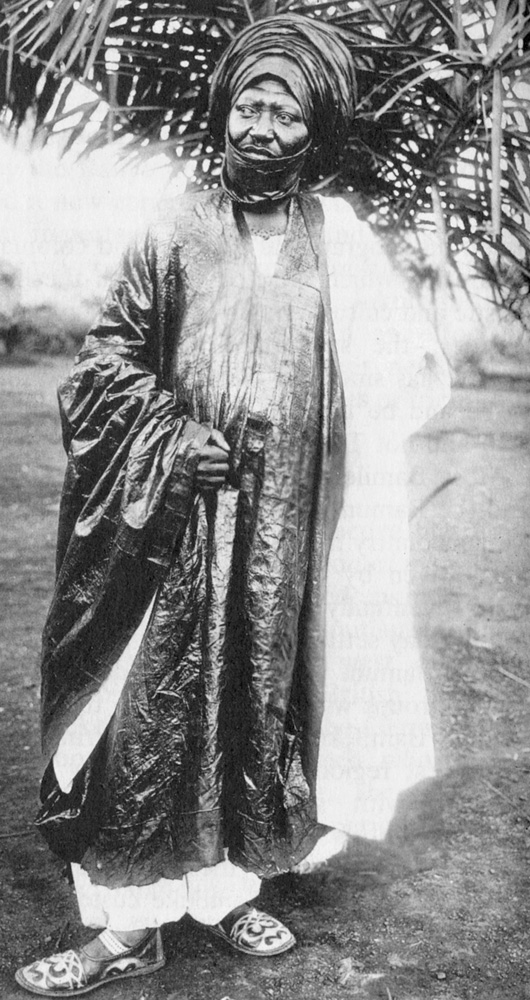 Czarno-białe zdjęcie przedstawia króla Ibrahima Njoyę. Mężczyzna stoi ubrany w ozdobną zwiewną szatę. W tle widać gałęzie egzotycznej rośliny.