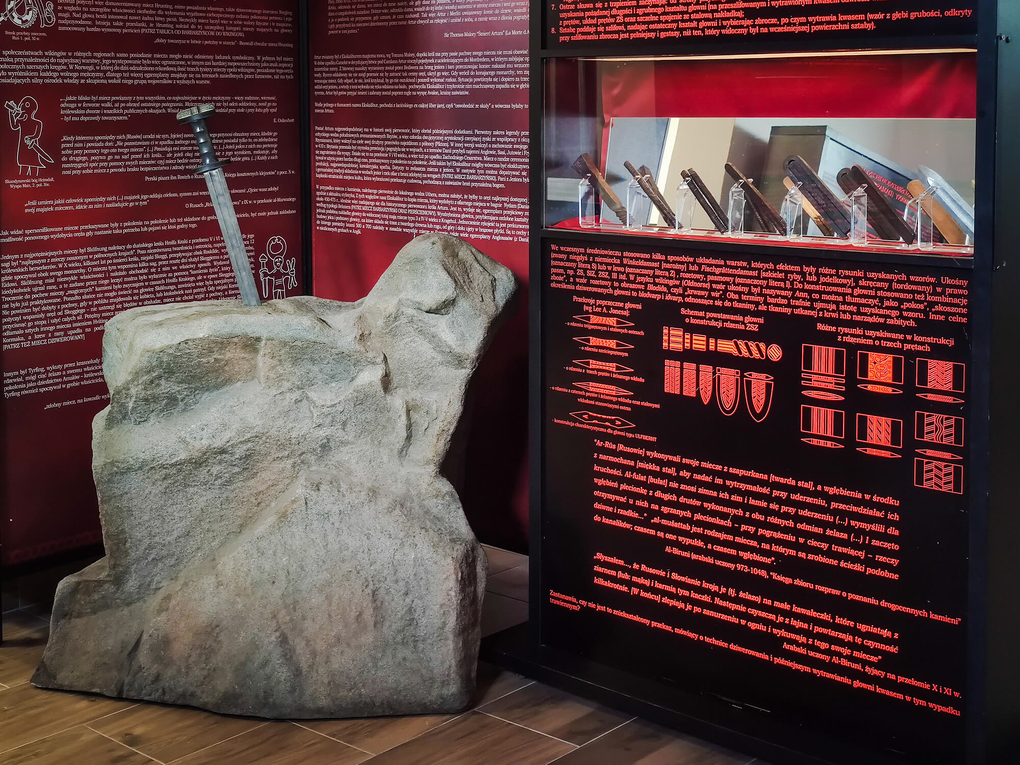 Fragment ekspozycji z instalacją "Ekskalibur". Miecz wbity w realistyczną imitację głazu. W tle plansze wystawowe, czarne z czerwonymi napisami. Po prawej szklana gablota z eksponatami: kawałkami ostrzy.