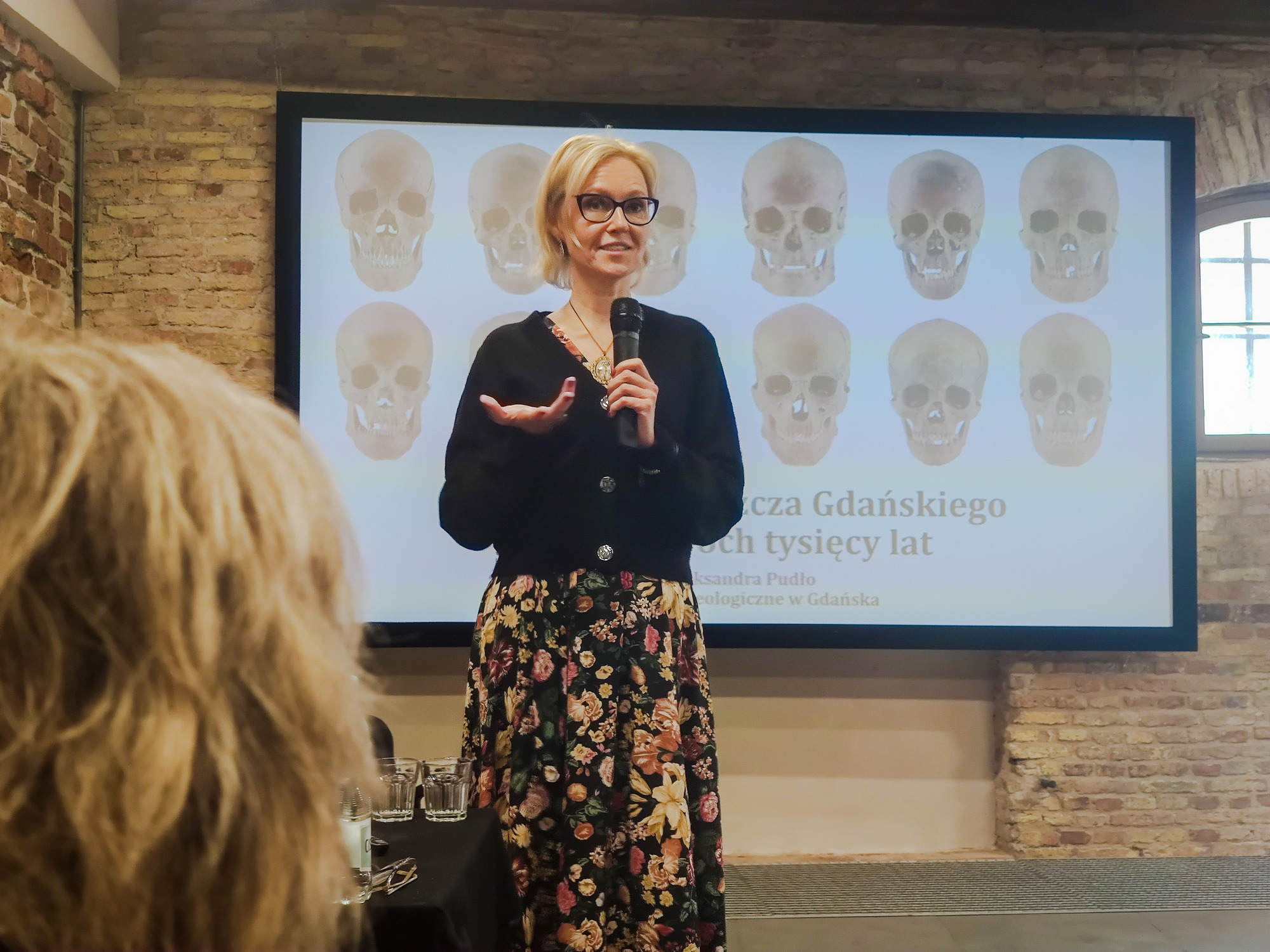 Kobieta w okularach stoi z mikrofonem w dłoni i przemawia. W tle widać ekran, na którym wyświetlone są zdjęcia ludzkich czaszek.